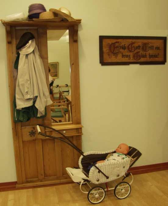 Garderobe mit Spiegel, Jacke, Hut, Bild an der Wand, davor alter Puppenwagen (Wird bei Klick vergrößert)