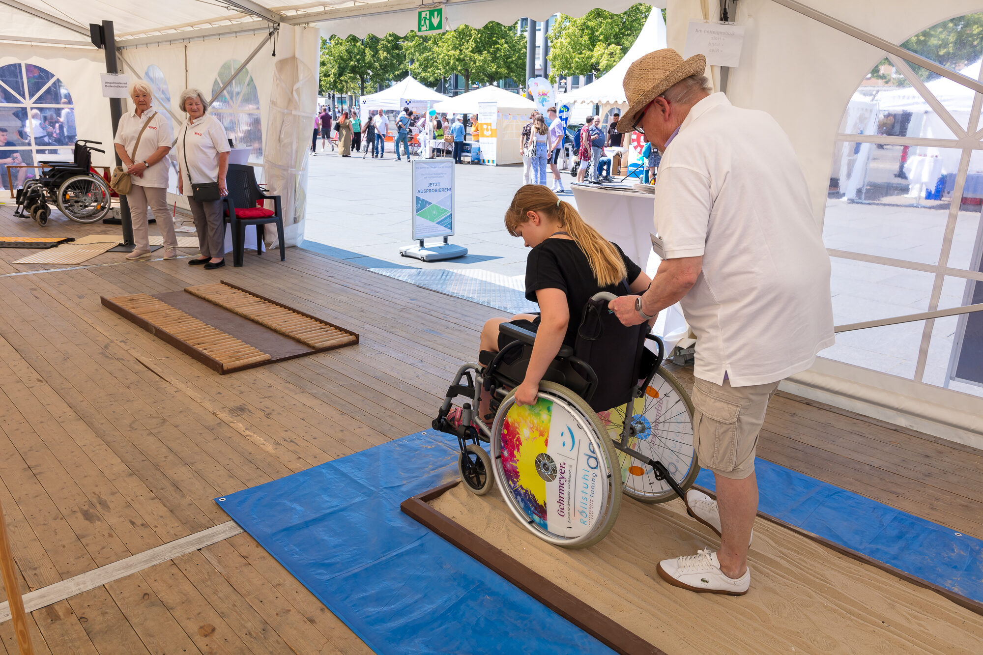 Impressionen Inklusionstage - Sensibilisierungsparcours, eine Rollstuhlfahrerin übt auf einem sandigen Untergrund mit Unterstützung (Wird bei Klick vergrößert)