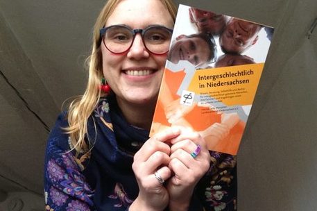 Eine Person mit langen Haaren und Brille hält auf dem Foto eine Broschüre Intergeschlechtlich in Niedersachsen