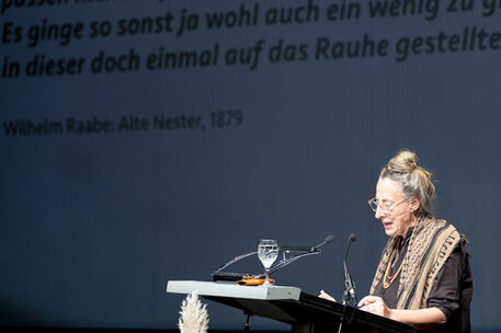 Gezeigt wird Judith Hermann, die gerade am Rednerpult ihre Rede hält. Sie trägt eine Brille, hat ihre Haare zu einem Dutt gebunden und trägt ein Halstuch
