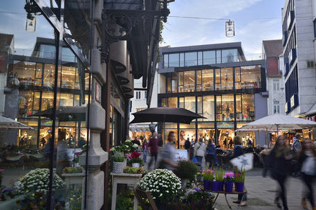 Das Bild zeigt die belebte Braunschweiger Fußgängerzone mit Menschen und aufgespannten Sonnenschirmen. Im Vordergrund ist ein Blumengeschäft zu sehen und im Hintergrund die beleuchtete Fassade eines Modegeschäfts.