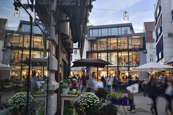 Das Bild zeigt die belebte Braunschweiger Fußgängerzone mit Menschen und aufgespannten Sonnenschirmen. Im Vordergrund ist ein Blumengeschäft zu sehen und im Hintergrund die beleuchtete Fassade eines Modegeschäfts. (Wird bei Klick vergrößert)