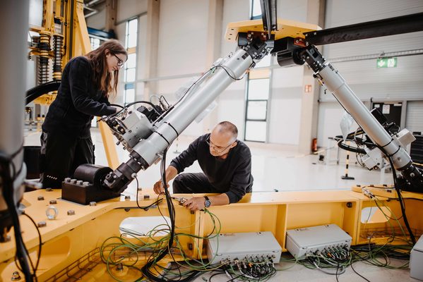 Das Bild zeigt einen knieenden Mann und eine stehende Frau, die an einer Maschine arbeiten. (Wird bei Klick vergrößert)