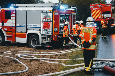 Die Feuerwehr Braunschweig bei einem Einsatz. Menschengruppe und Feuerwehreinsatzfahrzeug an einer Straße.