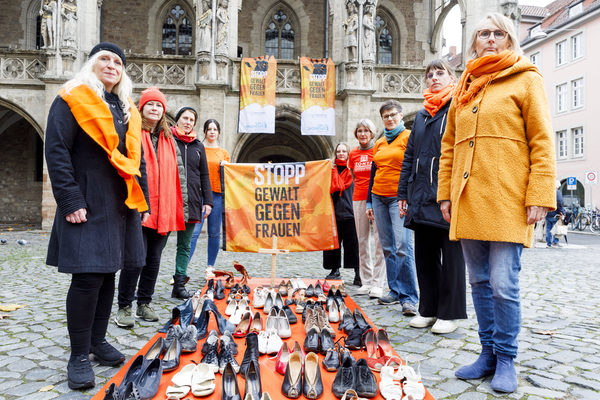 Frauen in oranger Kleidung stehen um eine orange Decke auf der 113 Paar Frauenschuhe stehen im Hintergrund eine Fahne mit den Worten Stopp Gewalt Gegen Frauen und dahinter das Altstadtrathaus in Braunschweig (Wird bei Klick vergrößert)