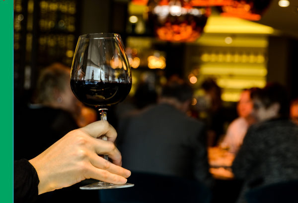 Frauenhand mit einem Glas Rotwein und eine Gruppe Menschen in einer Gaststätte im Hintergrund