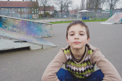 Ein Kind sitzt vorne im Bild. Im Hintergrund ist ein Skatepark zu sehen.