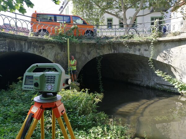 Ein Tachymeter steht am Ufer eines Flusses, im Hintergrund ist ein Brückenbauwerk. (Wird bei Klick vergrößert)