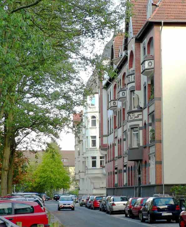 Entlang einer Straße in einem Wohngebiet mit Stadthäusern und parkenden Autos stehen Bäume.