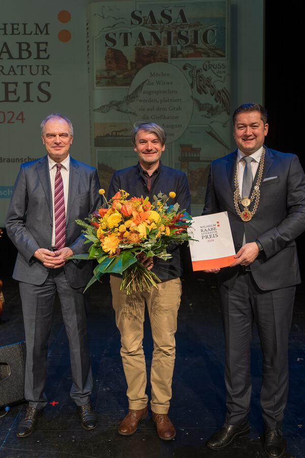 Foto des Preisträgers Sasa Stanisic (Mitte, mit gelben Blumenstrauß in den Händen), Intendant Stefan Raue (links) und Oberbürgermeister Thorsten Kornblum (rechts, überreicht dem Preisträger die Urkunde), im Hintergrund das Cover des mit dem Literaturpreis ausgezeichneten Buchs (Wird bei Klick vergrößert)