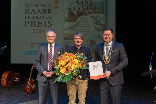 Foto des Preisträgers Sasa Stanisic (Mitte, mit gelbem Blumenstrauß in den Händen), Intendant Stefan Raue (links, von Deutschlandfunk) und Oberbürgermeister Torsten Kornblum (rechts, überreicht die Urkunde) (Wird bei Klick vergrößert)