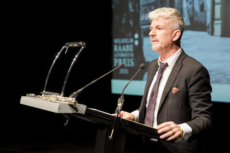 Ein Foto zeigt Heinz Strunk, Preisträger des Wilhelm Raabe-Literaturpreises 2016. Er steht am Rednerpult, trägt einen Anzug, richtet seinen Blick auf das Publikum und hat kurze grauweiße Haare
