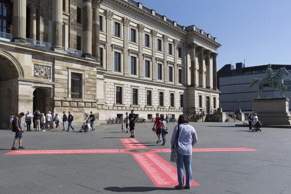 Auf dem Schlossplatz in Braunschweig klebt ein großes rotes Kreuz mit Namenslisten am Boden. Menschen bleiben stehen und schauen darauf. (Wird bei Klick vergrößert)