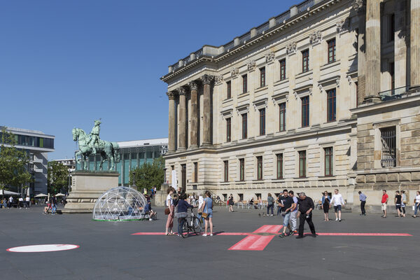 Auf dem Schlossplatz in Braunschweig klebt ein großes rotes Kreuz mit Namenslisten am Boden. Im Hintergrund ist eine durchsichtige Kuppel zu sehen. Menschen bleiben stehen und schauen auf das Kreuz am Boden. (Wird bei Klick vergrößert)