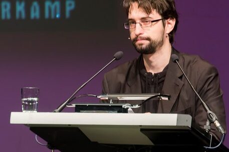 Gezeigt wird Clemens J. Setz, Preisträger des Wilhelm Raabe-Literaturpreises 2015. Er steht am Rednerpult mit Blick auf das Publikum, hat eine Brille, kurze braune Haare und einen kurzen Bart