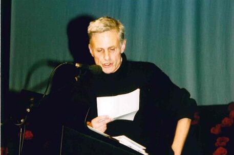 Der erste Wilhelm Raabe-Literaturpreis ging 2000 an Rainald Goetz. Hier sieht man ein Foto des Autors mit kurzen blonden Haaren, schwarz gekleidet vor dem Theatervorhang, wie er gerade von einem A4-Blatt liest.