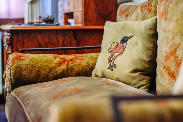 Sofa des Arbeitszimmers mit Blumenmuster vor der Schreibtischkante, auf dem Sofa ein Kissen mit einem gezeichneten Raben, der einen Anzug trägt (Wird bei Klick vergrößert)