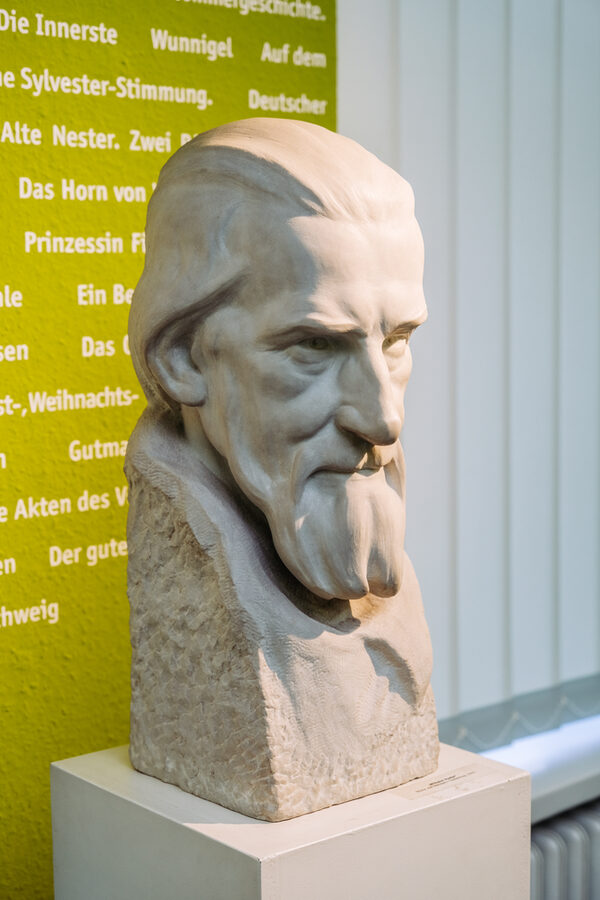 Büste von Wilhelm Raabe auf einem weißen Sockel, im Hintergrund ist ein abgeschnittener weißer Text auf grüner Wand sowie der weiße Vorhang am Fenster zu sehen (Wird bei Klick vergrößert)