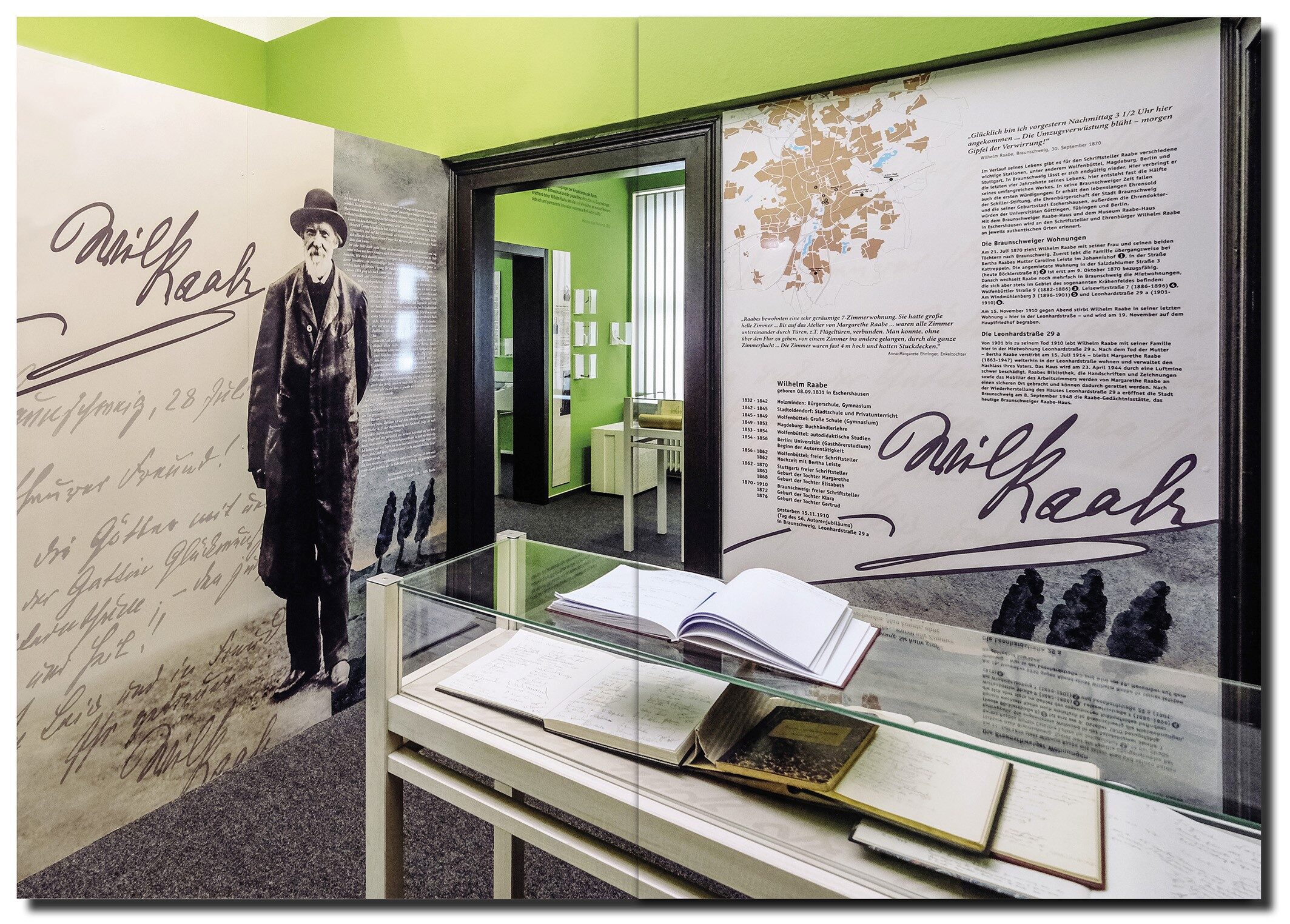 Eingangsbereich des Raabe-Hauses, im Vordergrund ist die Vitrine mit Gästebüchern zu sehen, dahinter Infotafeln zum Leben Wilhelm Raabes sowie die offenstehende Tür des Museums (Wird bei Klick vergrößert)