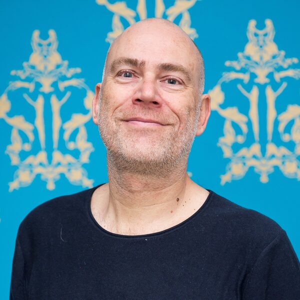Andreas Jäger vor der blauen Tapete mit Goldmuster vom Veranstaltungsraum des Raabe-Hauses, Glatze und graue Bartstoppeln, lächelt in die Kamera und trägt ein schwarzes T-Shirt (Wird bei Klick vergrößert)