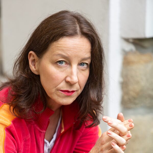 Verena Noll hat ihre Hände ineinander verhakt, hat schulterlanges leicht gelocktes Haar, trägt ein helles rotes Oberteil mit orangenen Streifen auf den Ärmeln (Wird bei Klick vergrößert)