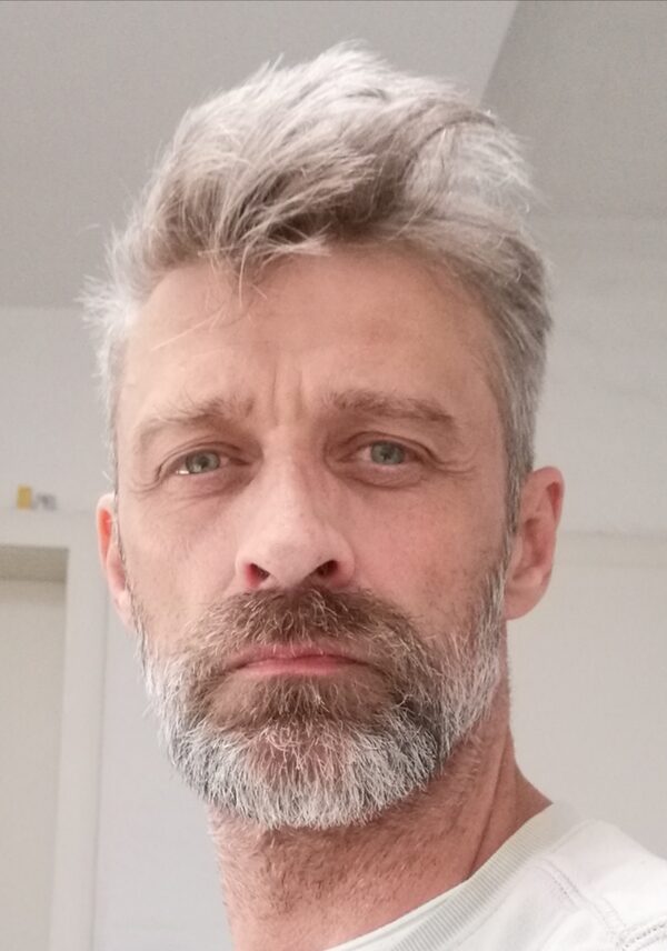 Dirk Lange, kurzer graubrauner Bart, etwas nach oben stehenden kurzen grauen Haaren mit Blick in die Kamera in einem weißen Raum (Wird bei Klick vergrößert)