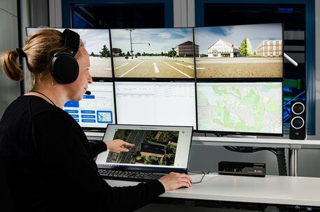 Das Bild zeigt eine Frau, die ein schwarzes Headset trägt, an einem Laptop. Hinter dem Laptop sind diverse Monitore zu sehen.